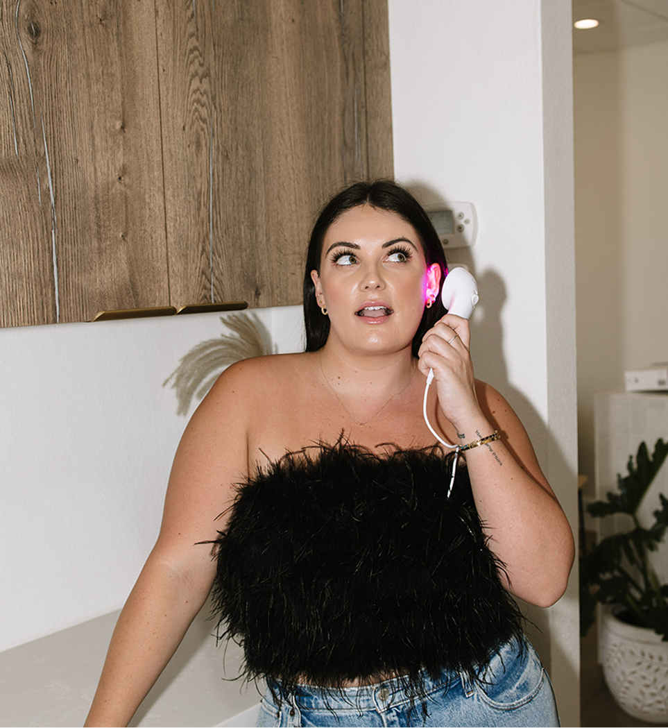 Woman wearing a black feathered top and a white phone with a pink light in a room.
